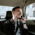 Smiling young man enjoying a phone call while seated in the back seat of a car during a daytime ride.